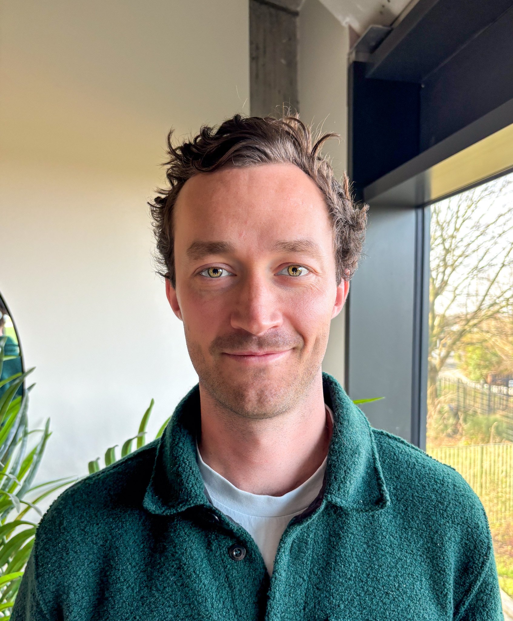 Will, one of the Cofounders of WebXecurity, smiling, wearing a green fleece jacket over a white shirt, standing near large windows with a view of trees outside.