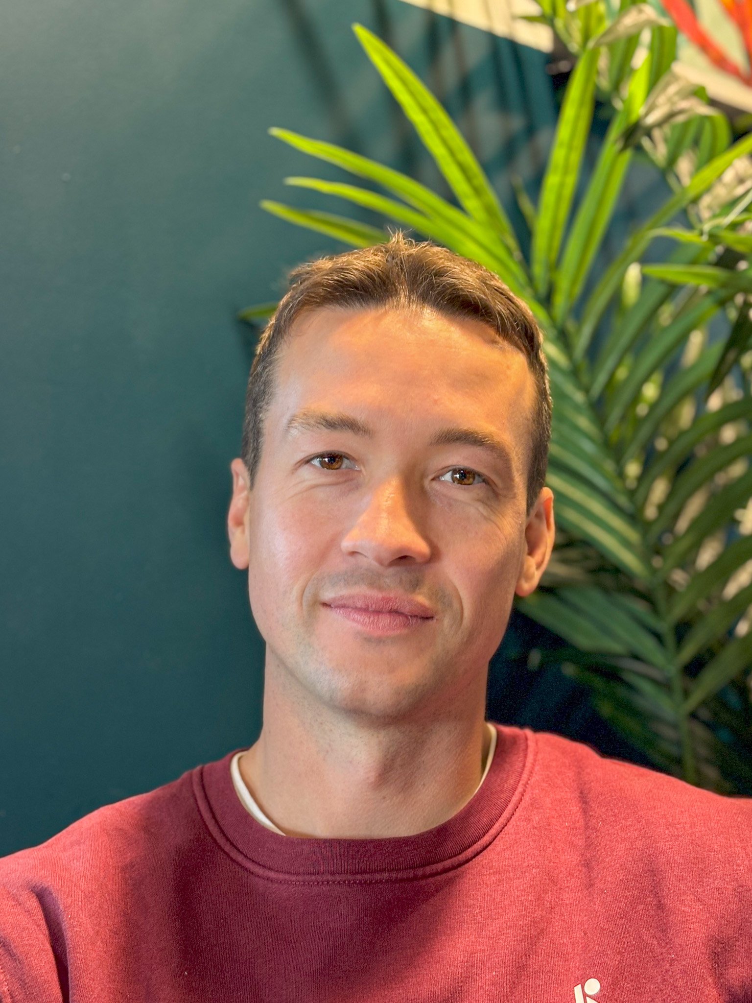 Stuart, one of the Cofounders of WebXecurity, smiling subtly, wearing a maroon sweatshirt, stands indoors in front of a dark green wall and a large green plant.